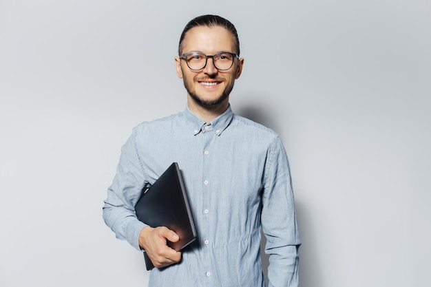 Retrato de estudio de un joven sonriente con un portátil en la mano sobre fondo blanco con camisa azul y anteojos