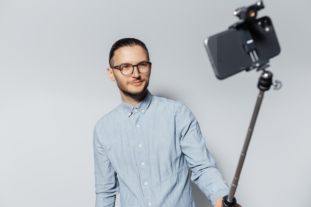 Retrato de estudio de un joven sonriente que toma una foto selfie a través de un teléfono inteligente y un palo Fondo gris claro Con anteojos y camiseta azul