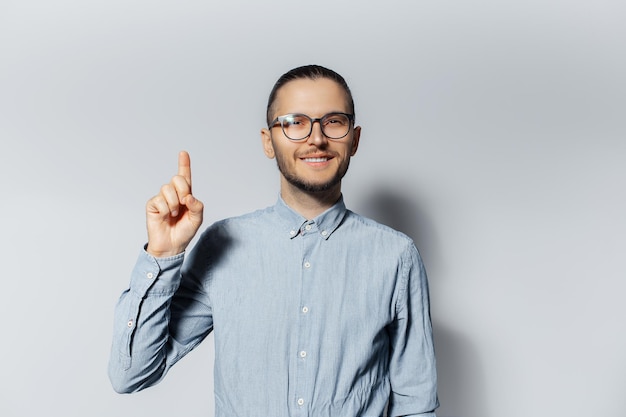 Retrato de estudio de un joven sonriente señalando con el dedo índice hacia arriba sobre fondo blanco usando anteojos y camisa azul Nuevo concepto de idea