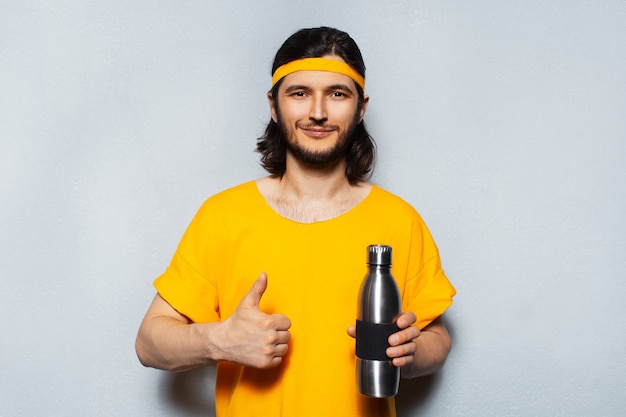 Foto retrato de estudio de joven sosteniendo una botella de agua termo de acero, mostrando los pulgares hacia arriba sobre el fondo de la pared con textura gris con banda amarilla para la cabeza y la camisa.