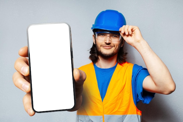 Retrato de estudio de un joven trabajador de la construcción sonriente que sostiene un gran teléfono inteligente con una pantalla en blanco en la mano que muestra un dispositivo cerca de la cámara con una maqueta sobre fondo blanco Con uniforme especial