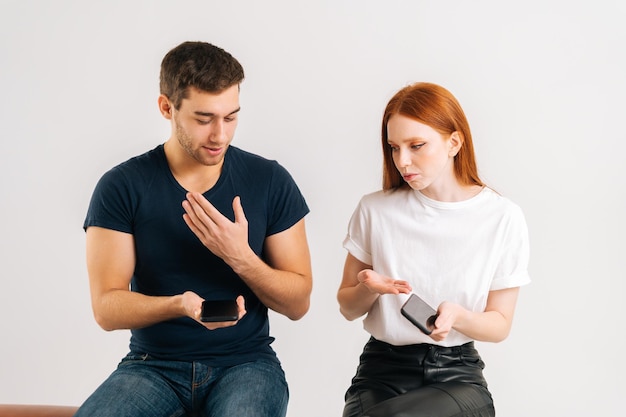 Retrato de estudio de una joven triste enojada con su novio usando un teléfono inteligente todo el tiempo sin prestarle atención en un fondo blanco aislado