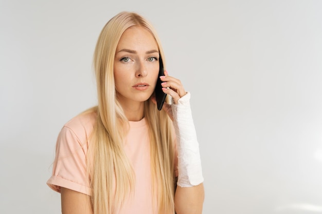 Retrato de estudio de una joven triste herida con un brazo roto envuelto en una venda de yeso hablando de un teléfono inteligente en un fondo blanco aislado mirando a la cámara