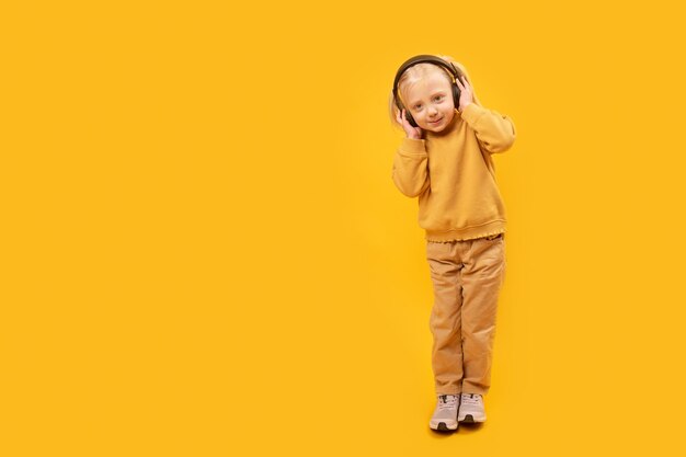 Retrato de estudio linda niña preescolar en traje amarillo escucha música con auriculares Crecimiento completo sobre fondo amarillo Maqueta de espacio de copia