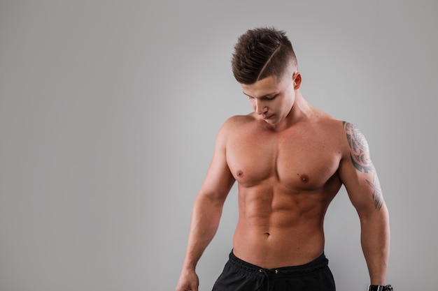 Retrato de estudio masculino de un apuesto joven culturista atlético modelo con cabello y un cuerpo sexy musculoso desnudo con un tatuaje en un fondo gris Modelo de hombre fuerte en estudio