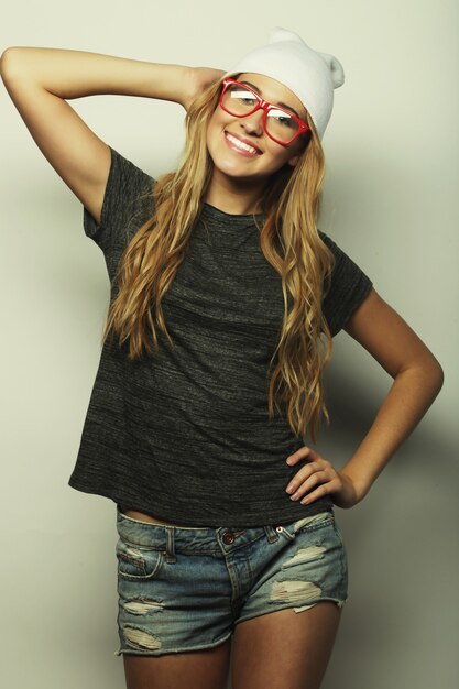 Retrato de estudio de moda de mujer rubia bastante joven inconformista con gafas, vistiendo elegante camiseta urbana y sombrero, sobre fondo blanco.