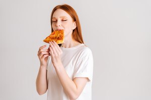 Foto retrato de estudio de mujer joven alegre con los ojos cerrados apetito comiendo deliciosa pizza de pie sobre fondo blanco aislado bonita mujer pelirroja comiendo comida sabrosa