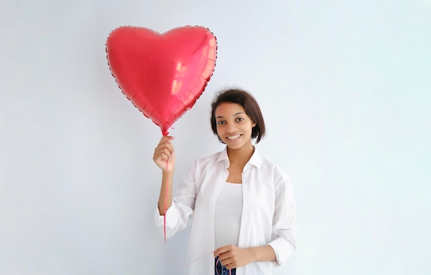 Retrato de estudio de mujer joven con piel oscura la pared roja festiva con globo en forma de corazón. Primer plano, fondo aislado, espacio de copia.