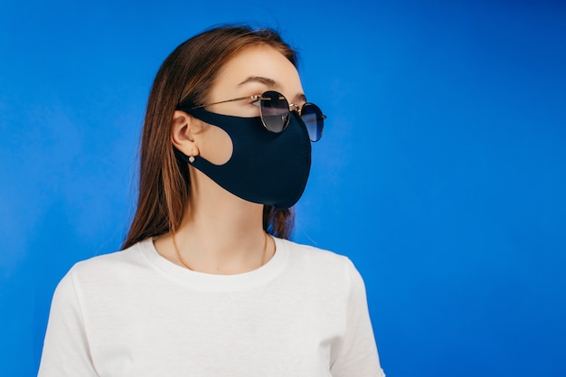 Retrato de estudio de una mujer joven que llevaba una máscara negra y gafas de sol, de cerca, aislado sobre fondo azul.