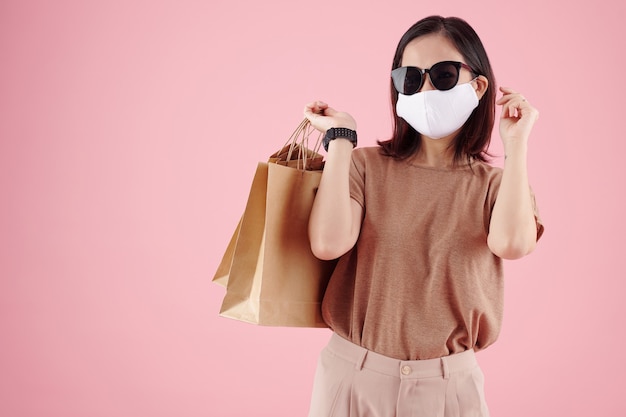 Retrato de estudio de mujer joven en ropa casual máscara médica y gafas de sol sosteniendo bolsas de la compra ...