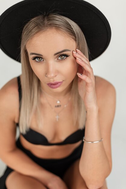 Retrato de estudio de mujer joven rubia de moda con increíbles ojos verdes en lencería de elegancia negra y sombrero mira a la cámara en el interior