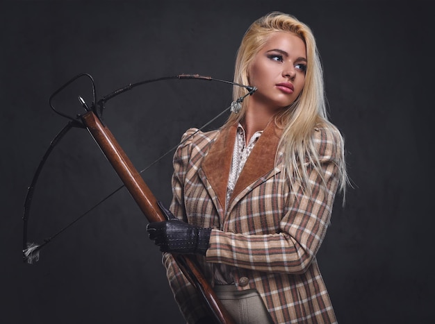 El retrato de estudio de una mujer rubia a la antigua sostiene una ballesta sobre un fondo gris.