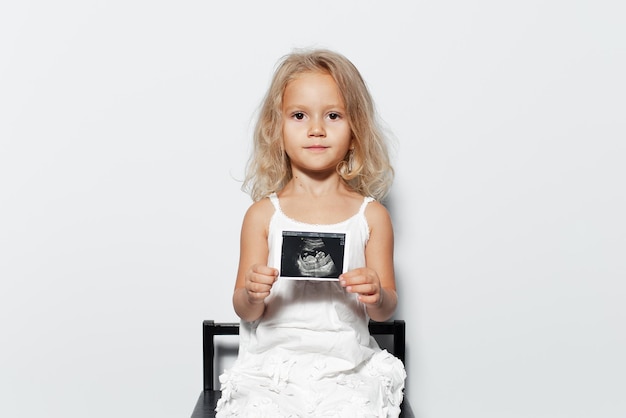 Retrato de estudio de niña con cabello rubio sosteniendo ecografía sobre fondo blanco.