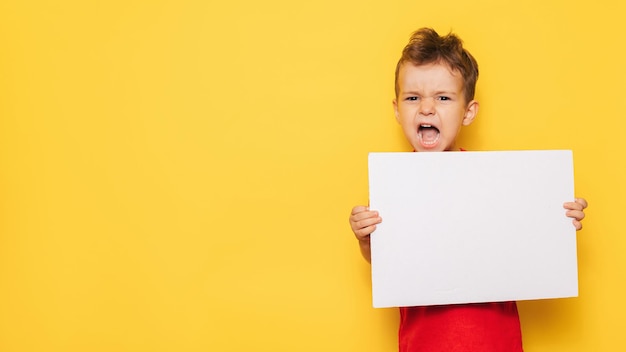 Retrato de estudio de un niño gritando con un cartel blanco limpio en sus manos sobre un fondo amarillo brillante, con un lugar para su texto o publicidad.