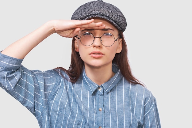 Retrato de estudio de primer plano de una joven caucásica posando para un anuncio con una gorra gris de camisa azul y anteojos transparentes redondos mirando hacia otro lado con la mano en la frente