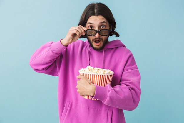 Retrato de un excitado joven barbudo morena hombre vestido con sudadera con capucha que se encuentran aisladas sobre la pared azul, comiendo palomitas de maíz, viendo películas en gafas 3 d