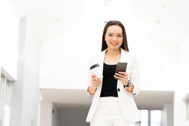 Retrato de una exitosa empresaria asiática de mediana edad sosteniendo un teléfono inteligente y un café sonriendo a la cámara