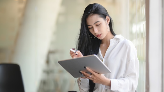Retrato de exitosa empresaria asiática con tableta digital mientras está de pie en la oficina moderna