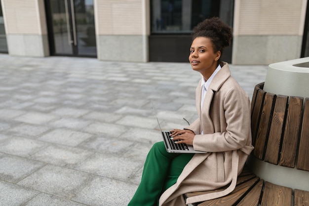 Retrato de una exitosa joven negra con una laptop en un banco cerca del centro de negocios