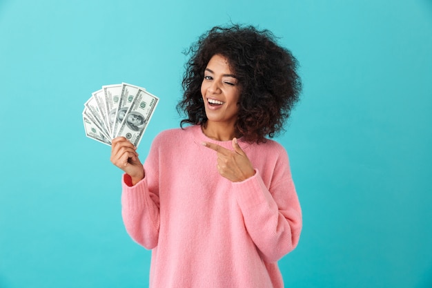 Retrato de la exitosa mujer estadounidense de 20 años con peinado afro sosteniendo un montón de billetes en dólares, aislado sobre la pared azul