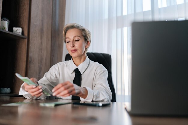 Retrato de una exitosa mujer de negocios de mediana edad contando dinero en el lugar de trabajo sentada a la mesa con