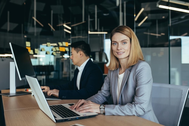 Retrato de una exitosa mujer de negocios miembro del equipo de negocios mirando la cámara y sonriendo trabajando en una laptop en una oficina moderna