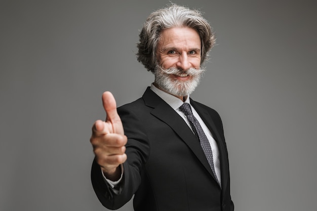 Retrato de exitoso empresario adulto vistiendo traje negro formal sonriendo y señalando con el dedo a la cámara aislada sobre pared gris