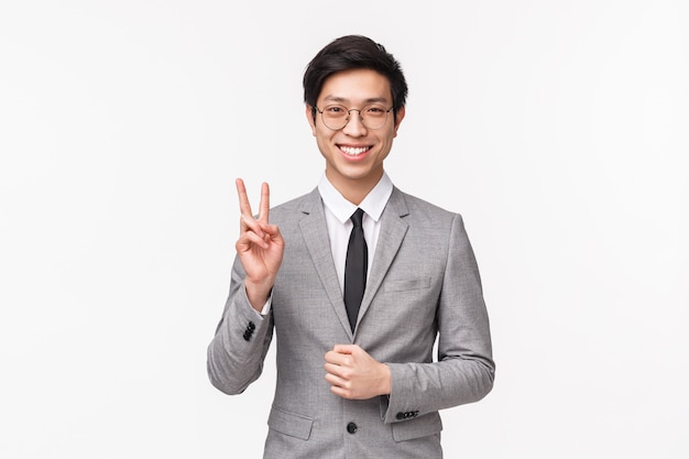 Retrato de exitoso empresario asiático joven feliz y despreocupado en traje formal gris, mostrando el signo de la paz y sonriendo, informar al equipo de la compañía que firmaron un acuerdo con éxito, en una pared blanca