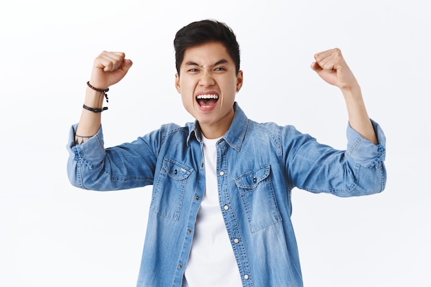 Retrato de un exitoso hombre asiático triunfante que bombea el puño regocijándose gritando sí hurra a la cámara ganando celebrando la victoria emocionado por el gol del equipo regocijándose por el fondo blanco