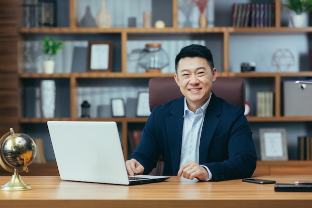 Retrato de un exitoso hombre de negocios asiático que trabaja en la oficina sentado en la mesa mirando la cámara y sonriendo feliz banquero con laptop