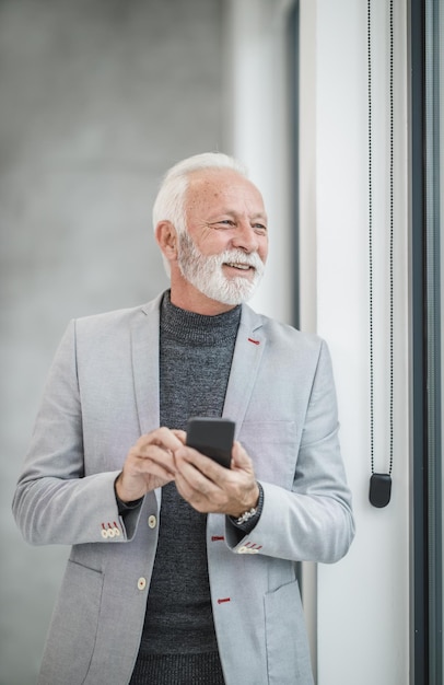 Retrato de un exitoso hombre de negocios senior que usa un teléfono inteligente mientras se encuentra en una oficina moderna y mira por la ventana.