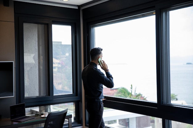 Retrato de un exitoso hombre de negocios con traje de pie usando un teléfono inteligente mirando por la ventana.