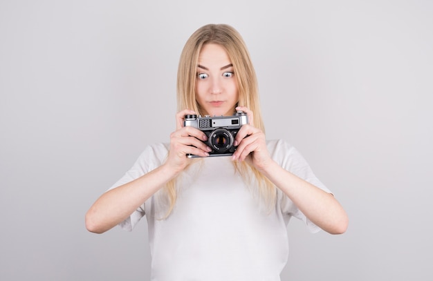 Retrato expressivo de uma bela jovem loira surpresa em uma camiseta branca com uma câmera nas mãos