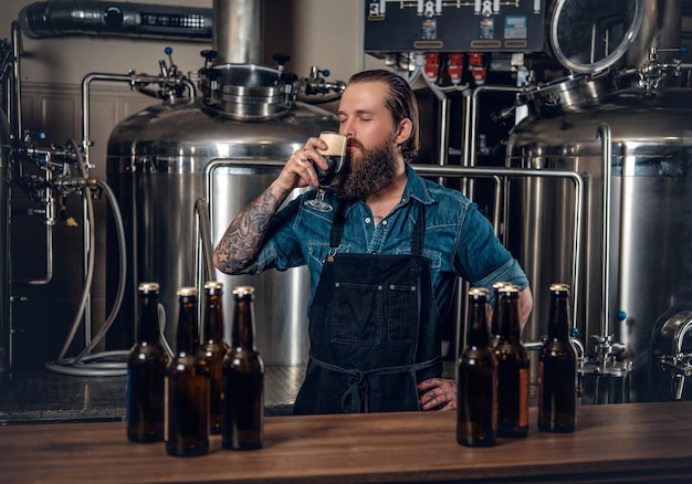 Retrato de un fabricante masculino tatuado y barbudo que prueba cerveza en la microcervecería.