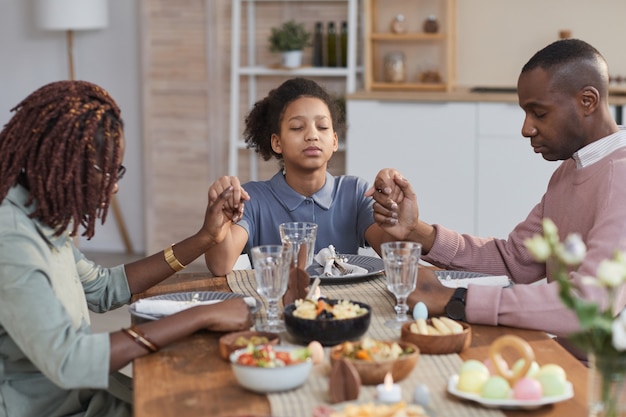 Retrato de familia afroamericana moderna diciendo gracia en la mesa de la cena en Pascua y tomados de la mano, espacio de copia