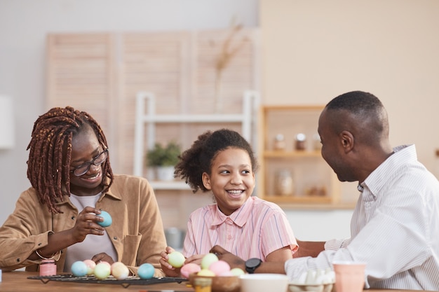 Retrato de familia afroamericana riendo pintando huevos de Pascua juntos mientras está sentado en la mesa de madera en el acogedor interior de una casa y disfruta del arte de bricolaje