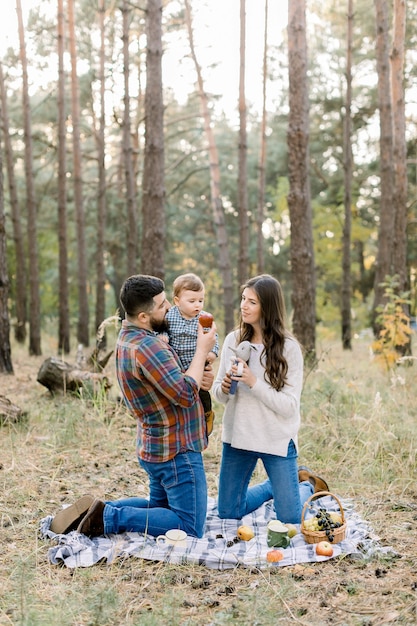 Retrato de familia al aire libre de padre alegre, madre y niño pequeño, divirtiéndose juntos, mientras está sentado en la manta en otoño hermoso bosque de pinos