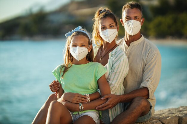 Retrato de una familia feliz abrazada con máscara protectora N95 pasando tiempo en la playa en la pandemia de la corona.