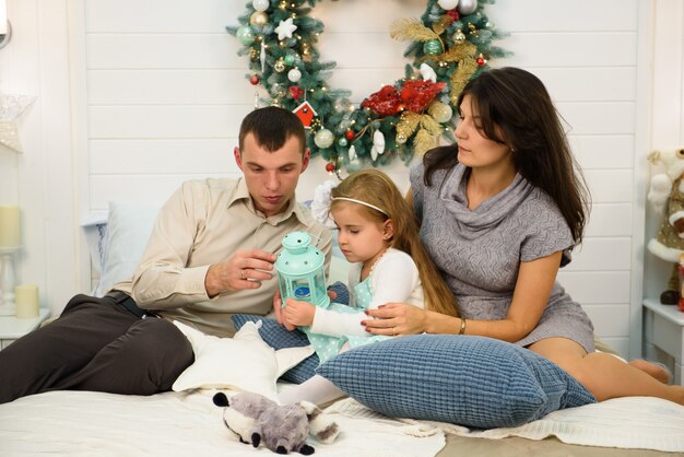 Retrato de familia feliz en Navidad, madre, padre e hijo sentados en la cama y encendiendo una vela en casa, decoración de chritmas a su alrededor