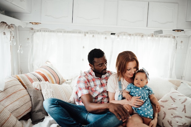 Retrato de una familia interracial feliz y sincera con una hija morena sentada en la cama de la caravana durante el día