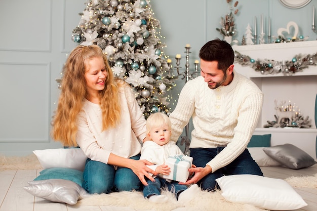 Retrato de una familia joven sentada en el suelo en la víspera de Navidad Hermosa madre con cabello largo y rizado y padre atractivo ayudando a su lindo hijo rubio a sostener una pequeña caja de regalo de Navidad