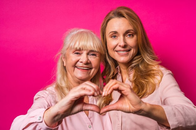Retrato de familia, mujer de mediana edad y madre senior formando un corazón con sus manos en la pared rosa