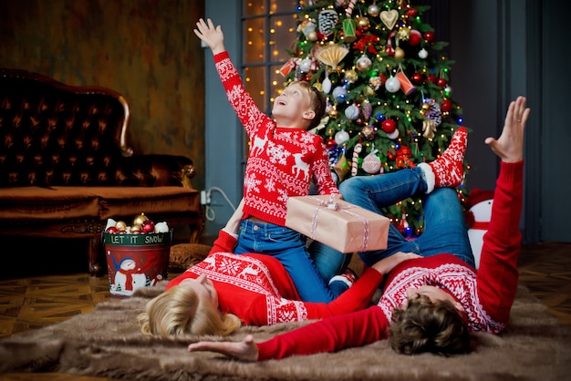 Retrato de familia de Navidad en suéteres tradicionales rojos en la sala de estar de vacaciones en casa, padres e hijos con caja de regalo.