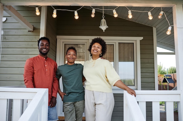 Retrato de una familia negra feliz parada en el porche de una casa nueva y sonriendo al espacio de copia de la cámara