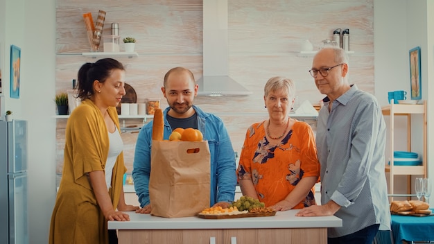 Retrato de familia video de cámara lenta sentado en la cocina moderna. Gente feliz sonriendo a la cámara en el comedor alrededor de la bolsa de papel con comestibles mirando a la cámara