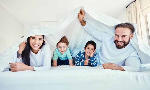 Foto retrato familiar y fortaleza de mantas en la cama con niños y padres felices y jugando en su casa cara debajo y sábana de niños con mamá y papá en el dormitorio divirtiéndose y despertándose, relájese y sonría