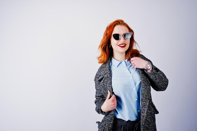 Retrato de una fantástica chica pelirroja con camisa azul y abrigo gris posando con gafas de sol en el estudio