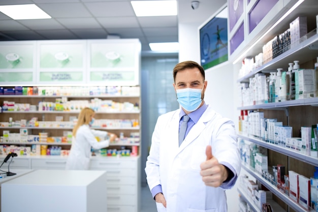 Retrato de farmacéutico con mascarilla y bata blanca que muestra los pulgares hacia arriba en la farmacia durante la pandemia del virus corona.