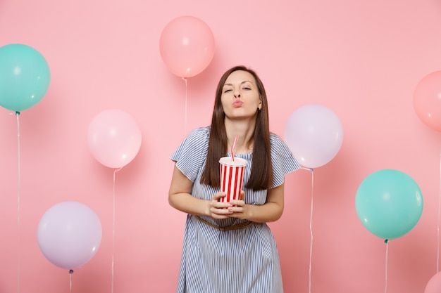 Retrato de fascinante mujer joven feliz con vestido azul que sopla labios beso sosteniendo un vaso de plástico de cola o soda sobre fondo rosa pastel con coloridos globos aerostáticos. Concepto de fiesta de cumpleaños.