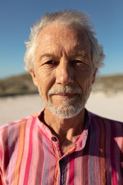 Retrato fechado de um homem caucasiano sênior na praia ao sol, vestindo uma camisa listrada, olhando para a câmera e sorrindo, contra um céu azul